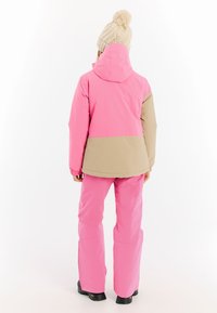 Rosa und beige wasserdichte Skijacke mit Kapuze, elastischen Bündchen und passenden rosa Hosen. Verfügt über ein einfaches Design und robuste schwarze Stiefel.