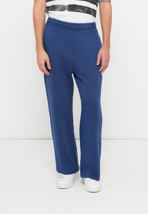 Blaue Sweatpants mit elastischem Bund, lockerem Schnitt und einfarbiger Textur. Kombiniert mit weißen Turnschuhen und einem gestreiften Hemd.