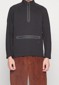 Pullover negro de manga larga con cuello alto y media cremallera; cuenta con un bolsillo con cremallera horizontal en la parte delantera y una tela texturizada.