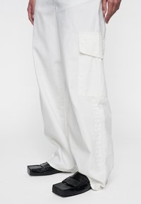 Witte cargobroek met een zijzak, voorzien van subtiele cijfers die langs het been zijn gedrukt. Gecombineerd met zwarte, chunky schoenen.