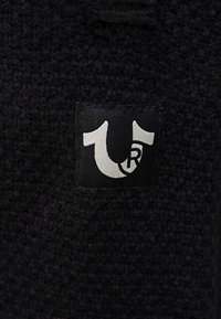 Czarna, teksturowana tkanina z małą czarną metką z białym logo, składającym się z stylizowanych liter "U" i "R" splecionych ze sobą.