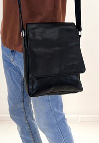 Borsa a tracolla in pelle nera con chiusura a pattina, caratterizzata da una superficie testurizzata e una tracolla regolabile. Grande tasca frontale per lo stivaggio.