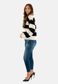 Maglione a righe bianche e nere in tessuto morbido, abbinato a jeans blu aderenti e scarpe col tacco nere, mostrato da un angolo laterale.