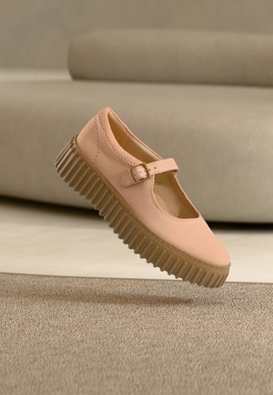 Chaussure Mary Jane rose clair avec semelle épaisse marron rainurée et bride à boucle, positionnée au-dessus d'un tapis beige et d'un fond de canapé.
