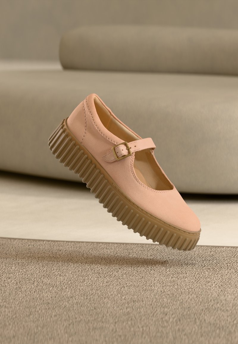 Chaussure Mary Jane rose clair avec semelle épaisse marron rainurée et bride à boucle, positionnée au-dessus d'un tapis beige et d'un fond de canapé.