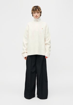 Asmuo, dėvintis baltą aukštu kaklu megztinį ir plačias juodas plataus kirpimo kelnes, stovintis priešais paprastą šviesų foną.