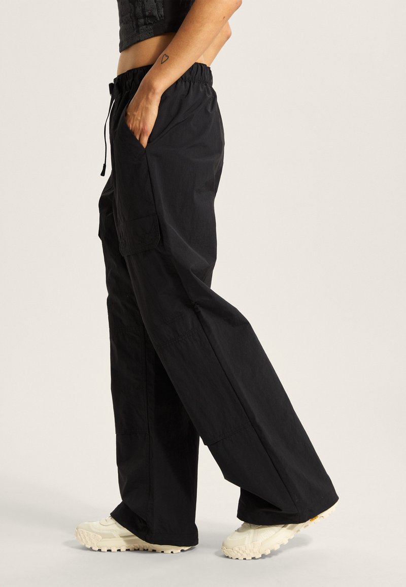 Pantalones cargo negros con un ajuste holgado, cinturilla elástica, bolsillos laterales y un tejido texturizado. Combinados con zapatillas chunky de color claro.