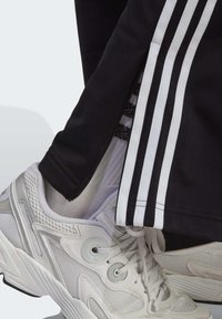 Svarta träningsbyxor med vita sidostråk, kombinerade med vita sportskor med nät och släta detaljer. Texturerad och funktionell design.