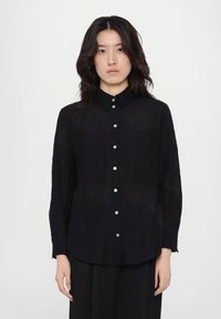 Zwarte, lange mouwen shirt van lichtgewicht stof, met een knopenlijst aan de voorkant, puntige kraag en twee borstzakken.