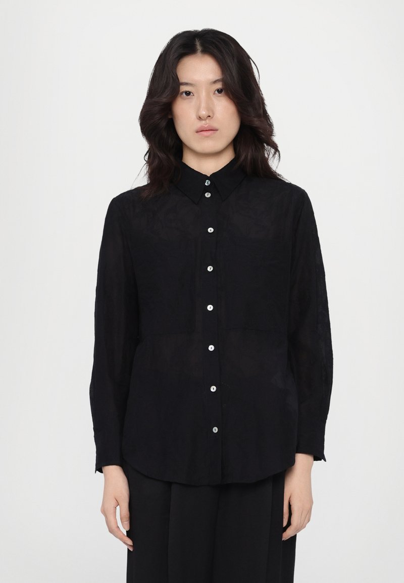 Zwarte, lange mouwen shirt van lichtgewicht stof, met een knopenlijst aan de voorkant, puntige kraag en twee borstzakken.