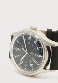 Montre-bracelet en acier inoxydable argenté avec un cadran noir, des chiffres arabes blancs, des aiguilles lumineuses, un bracelet en cuir noir, affichant l'heure 10:57:38.