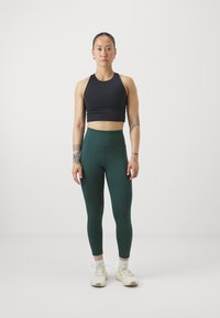 Čierny crop top a tmavozelené legíny so zvýšeným pásom, vyrobené zo strečového materiálu. Biely športový obuv s šnúrkami, postavený na jednoduchej pozadí.