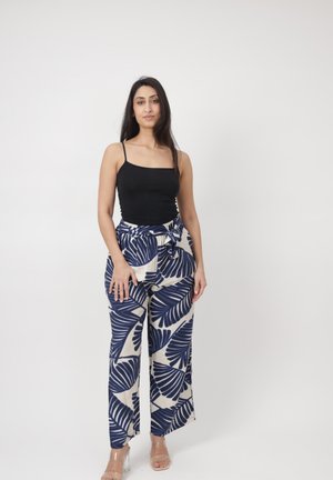 Kobieta stojąca na białym tle, ubrana w czarny top bez rękawów oraz beżowe spodnie z szerokimi nogawkami i dużym niebieskim wzorem liści, nosząca przejrzyste szpilki.