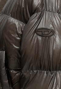 Veste matelassée noire brillante en gros plan montrant la texture du tissu, les coutures, la fermeture éclair et le patch logo brodé noir ovale sur la manche.