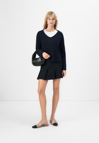 Femeie îmbrăcată cu un pulover bleumarin peste un top alb, o fustă mini plisată neagră, pantofi slingback bicolori și ținând o geantă de piele neagră.