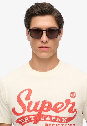 Gafas de sol negras con montura rectangular, usadas con una camiseta crema que lleva un texto rojo intenso "Super Dry Japan" y un emblema de marca registrada.