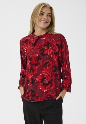 Smiling woman with blonde hair wearing a red floral long-sleeve blouse and black pants, hands in pockets, against a plain background.