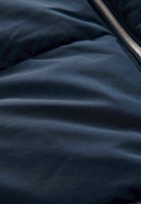 Tissu d'une veste matelassée bleu marine foncé avec une texture lisse et brillante. Présente des plis subtils et une fermeture éclair métallique le long d'un bord.