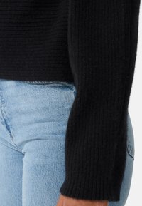 Pull noir côtelé au toucher doux, doté de manches longues et d'une coupe ajustée, porté sur un jean en denim bleu clair.