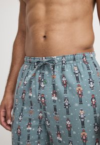 Pantalones cortos de pijama turquesa con patrón de cascanueces. Hechos de una tela suave, cuentan con una cintura elástica, cordón y un detalle de botón verde.