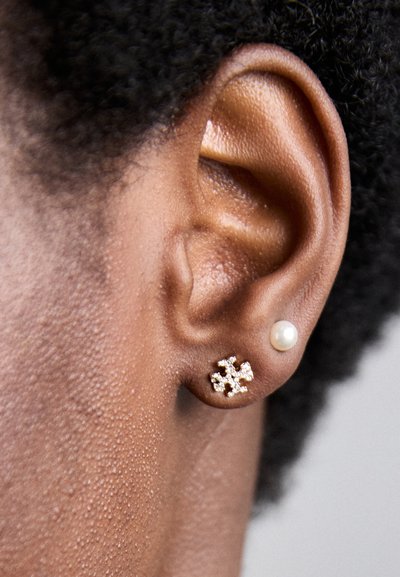Boucle d'oreille en forme de flocon de neige doré avec des accents scintillants, associée à une puce en perle blanche ronde, présentée sur une peau texturée.