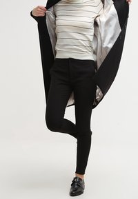 Černý dlouhý kabát, který odhaluje bílý pruhovaný svetr uvnitř, v kombinaci s černými přiléhavými kalhotami a černými botami na přezku.