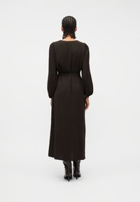 Γυναίκα με κοτσίδες, με την πλάτη γυρισμένη, φοράει ένα μακρυμάνικο, σκουροκάστανο μίντι φόρεμα και μαύρες μπότες με τακούνια σε λευκό φόντο.