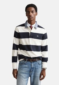 Tamsiai mėlynas ir baltas dryžuotas ilgomis rankovėmis polo marškinėliai su džinsiniu apykakle, derinami su šviesiai mėlynais džinsais ir rudu diržu, su mažyčiu logotipu.