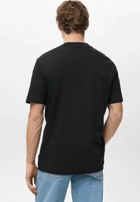 Moški s kodrasto frizuro, oblečen v preprosto črno majico s kratkimi rokavi in svetlo modre kavbojke, obrača hrbet proti enobarvni beli podlagi.