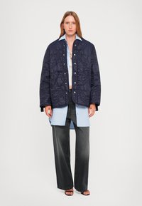 Navy quilted jacket with textured pattern, silver snap buttons, layered over a light blue shirt and black wide-leg pants. Black sandals.