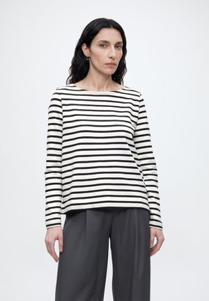 Femme aux longs cheveux foncés portant un t-shirt à manches longues rayé noir et blanc et un pantalon ample gris foncé, debout sur un fond blanc uni.