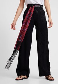 Pantalon noir à jambes larges avec un motif texturé et une ceinture foulard rouge contrastante ornée de motifs graphiques et de détails à franges. Des sandales complètent le look.
