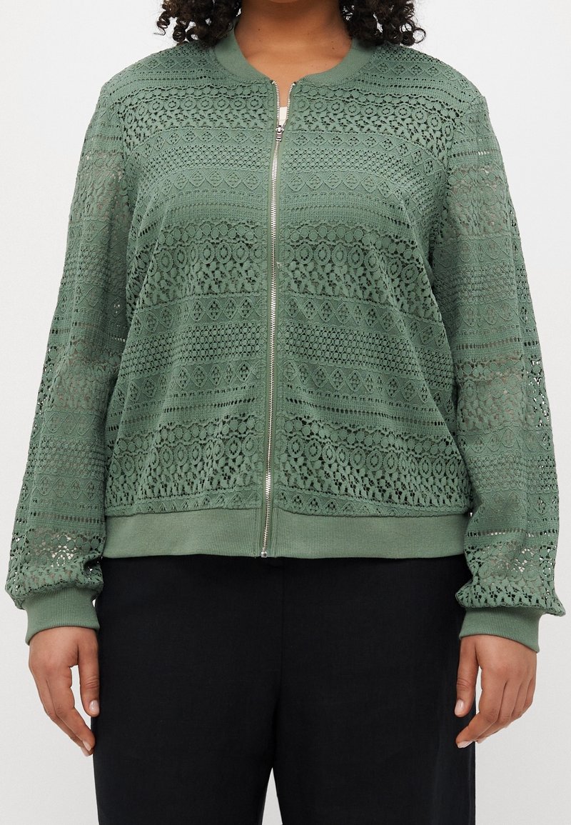 Femme portant une veste zippée en dentelle verte avec des motifs floraux et géométriques, associée à un pantalon noir sur fond blanc.