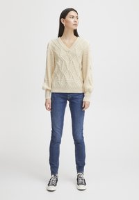 Pull crème en tricot torsadé avec un col en V, des motifs en relief et des manches ballons, associé à un jean slim fit bleu foncé et des baskets noires.