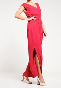 Robe longue rouge en tissu léger, avec des manches courtes, un décolleté en V profond et une fente sur le côté. Associée à des talons de couleur claire.