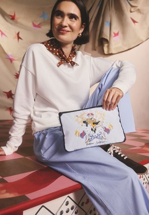 Geantă din pânză albastru deschis cu ilustrație grafică, margine neagră și accente multicolore. Model purtând un top alb și pantaloni albastru deschis, așezat pe o suprafață cu model.