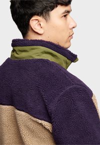 Fleece pullover with purple, olive green, and beige sections. Features a high collar and textured surface for warmth and comfort.