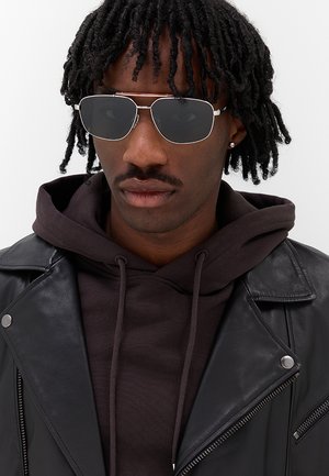 Giovane uomo con dread corti che indossa occhiali da sole aviator scuri, giacca nera in pelle e felpa scura con cappuccio davanti a uno sfondo semplice.