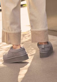 Grå ruskinds slip-on sko med en tyk gummisål og synlige syninger, båret med lyse, opsmøgede bukser på et fortov.