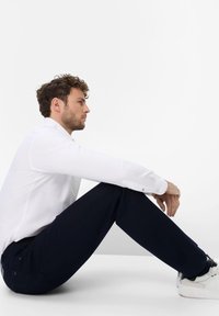 Camisa blanca de manga larga con botones en los puños, combinada con pantalones oscuros de color azul marino. Modelo sentado con zapatillas blancas, mostrando un estilo y un corte relajado.