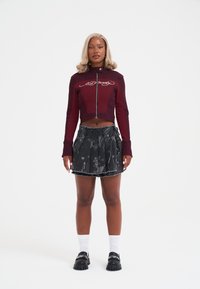 Kobieta stoi w maroonowej, krótkiej kurtce na zamek, czarnej spódniczce mini z nadrukiem, białych skarpetkach i czarnych butach na tle białym.