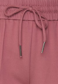 Rosa joggers med dragsko i midjan och metalliska ändar. Slät textur, avslappnad passform och sydda detaljer längs midjan.