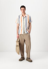Camisa de manga curta às riscas em cores pastéis (azul, laranja, verde) com um bolso, combinada com calças largas em bege e sapatos castanhos.