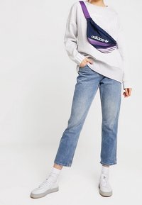 Šedá oversized mikina, modré džínové kalhoty, bílé tenisky a tmavě modrá a fialová ledvinka Adidas s logem. Outfit má ležérní styl.