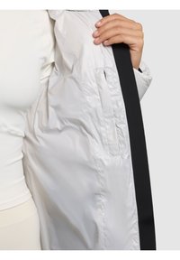 Ruka drži otvorenu bijelu jaknu s crnim rubom, pokazujući glatku unutarnju podstavu i unutarnji džep.