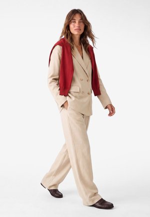 Mujer caminando con un traje cruzado beige con pantalones amplios, zapatos marrón oscuro y un jersey rojo sobre los hombros.