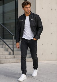 Young man walking outdoors wearing a black leather jacket, white shirt, black jeans, and white sneakers near concrete steps and wall.