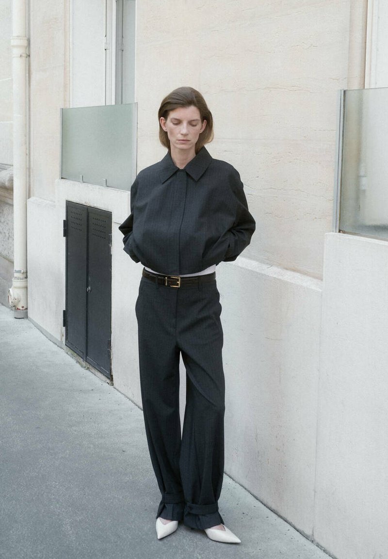 Femme en costume noir ample, les mains dans les poches, debout contre un mur en pierre claire sur un trottoir en ville, portant des chaussures blanches pointues.