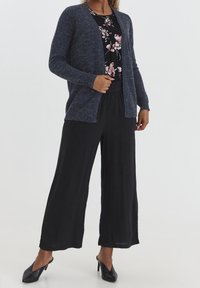 Žena oblečená v tmavě šedém kardiganu přes květinovou blůzu, černých širokých kalhotách a černých muly na podpatku, stojící na jednobarevném pozadí.