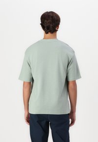 Homme portant une chemise vert clair à manches courtes et un pantalon sombre, debout de dos sur un fond blanc uni.